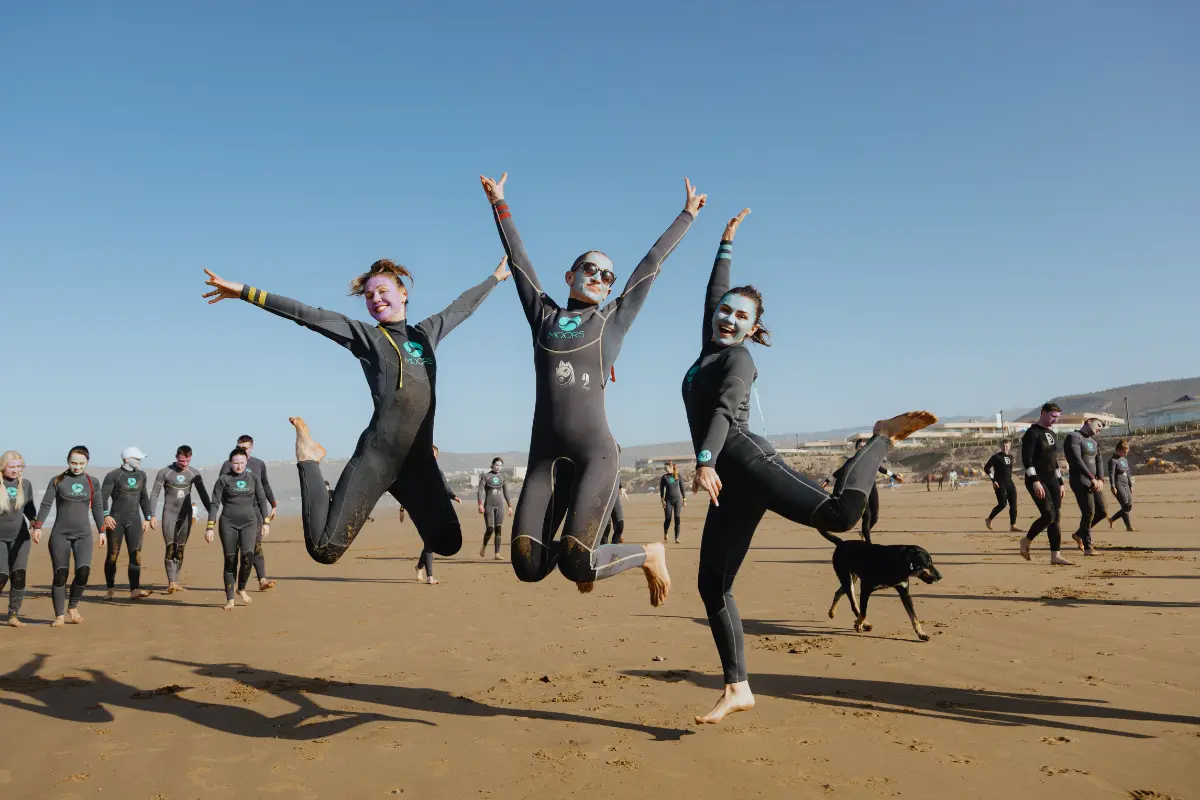 Three happy surfers jumping on the beach during a fun surf camp trip in Morocco, featuring a lively group and a dog.