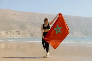 Young woman celebrating her surf trip to Morocco, holding the national flag on a beautiful beachfront. Represents the spirit of a surf camp Morocco experience in popular areas like Taghazout or Tamraght. Ideal for showcasing surf lessons, surf holidays, and surf camp packages.