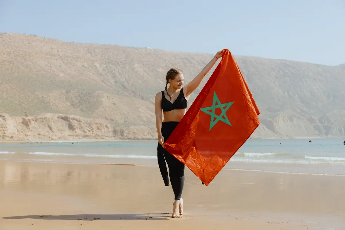 A smiling person holds a vibrant Moroccan flag on a wide, sunlit beach with hills in the background, representing a joyful surf holiday in Morocco.
