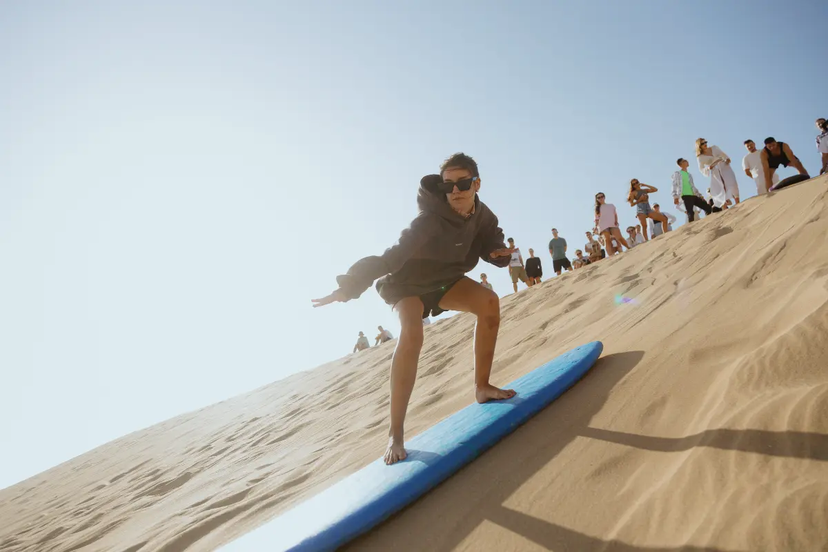 Young person sandboarding down a large sand dune with a blue board resembling a surfboard, while a group watches. Represents exciting activities included in a surf camp package in Morocco, near Taghazout or Tamraght, part of a fun surf trip.