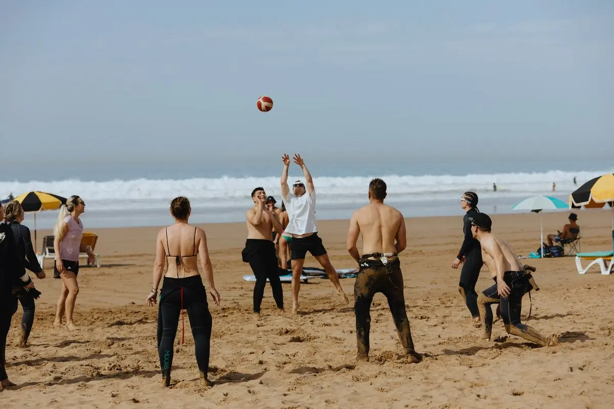 Guests enjoy beach volleyball, a fun activity offered at a surfcamp in Taghazout, Morocco. People in wetsuits and swimwear are playing on the sand with waves in the background, showing the lively atmosphere at this surf camp.