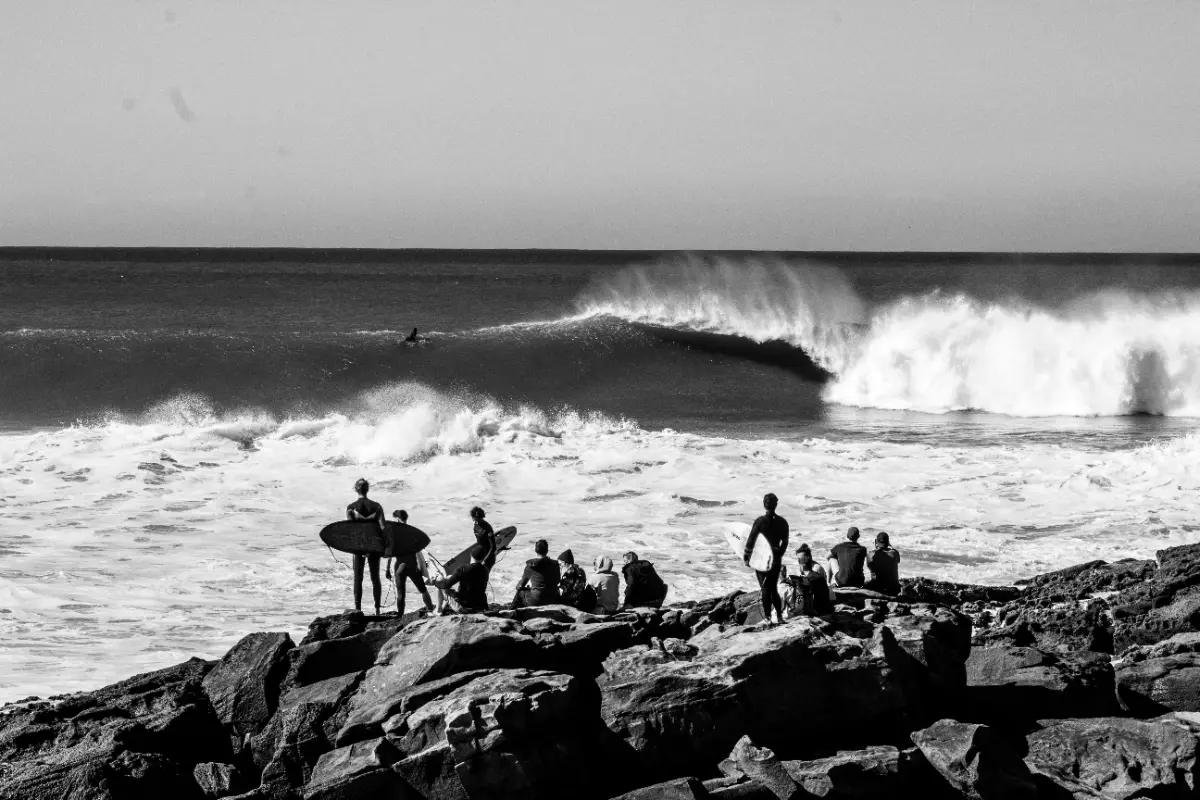 Surfers and onlookers sit on rocky shore observing a large wave breaking. A common sight near a surfcamp Taghazout, Morocco, known for its excellent surfing conditions.