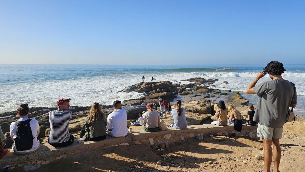 Besøkende sitter på muren ved Anchor Point og ser på surfingen i Taghazout