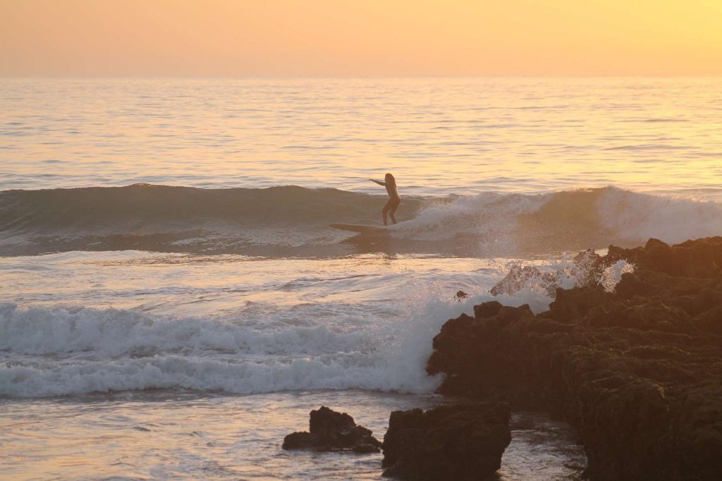 Surfer på en bølge i solnedgang ved steinene på Devil's Rock, Tamraght
