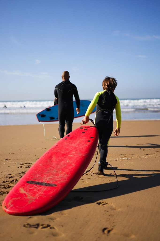 Far og søn bærer et rødt surfboard ud mod bølgerne på surfferie med familien i Marokko