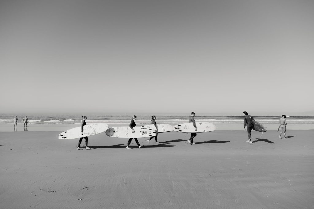 Foto en blanco y negro de alumnos de surf llevando sus tablas por la playa de Tamraght